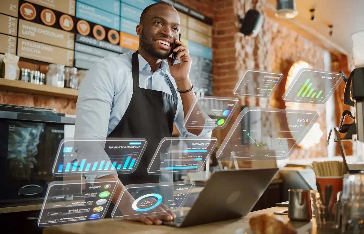 A cafe worker talks on a phone beside a laptop as digital charts float over the counter.